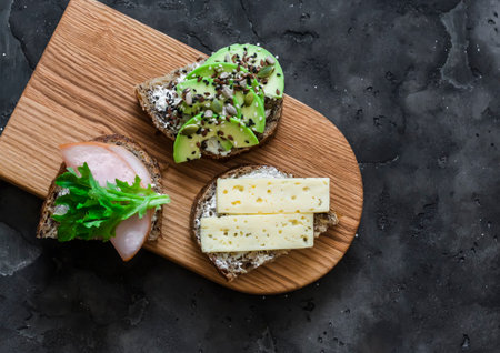 Breakfast, snack, tapas sandwiches - with avocado, ham, cheese on a wooden cutting board on a dark background, top viewの写真素材