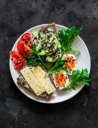 Sandwiches with avocado, cheese, seeds, boiled egg, tomato and green salad - delicious breakfast, snack on a dark background, top viewの写真素材