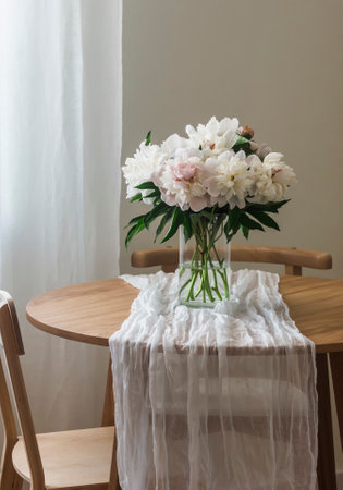 A bouquet of beautiful white peonies in a glass vase on a wooden round table with a white tableclothの写真素材