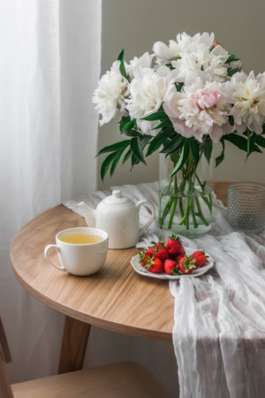 A bouquet of white peonies in a glass vase, chamomile tea, strawberries on a white round table in a cozy living roomの写真素材
