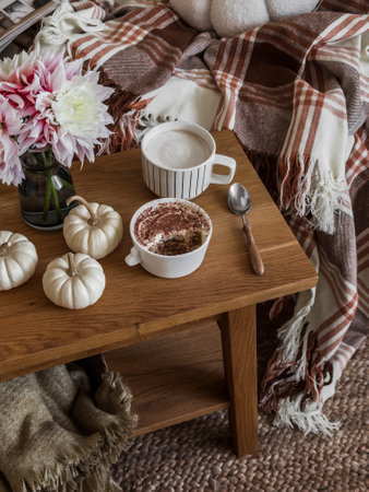 Coffee break - cappuccino, tiramisu, autumn decor on a wooden bench near the sofa in a cozy living roomの写真素材