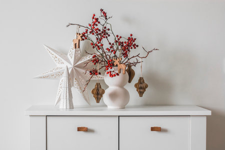Christmas composition on a white chest of drawers - a Christmas bouquet with cranberry branches and paper decorations, a Christmas star and a ceramic Christmas treeの写真素材