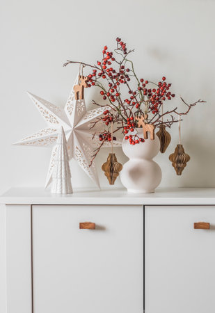 Scandinavian-style Christmas composition on a white chest of drawers - a Christmas bouquet with cranberry branches and paper decorations, a Christmas star and a ceramic Christmas treeの写真素材