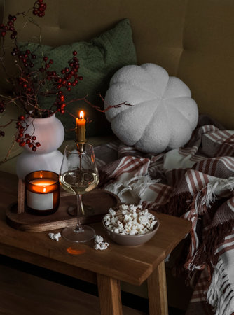 A glass of white wine, a bowl of popcorn, a lit candle on a wooden table next to the sofa in the evening living roomの写真素材