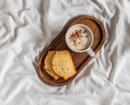 Slice of pound cake, a cup of cappuccino on a wooden tray on the white sheets of the bed. Breakfast in bedの写真素材