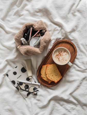 Aesthetics of the morning - cosmetic bag, planner notepad, coffee and cupcake on white sheets in bed. Cozy morningの写真素材