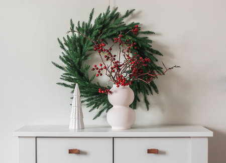 Scandinavian-style Christmas decor in the living room - a spruce wreath on the wall, a vase with cranberry branches on a white chest of drawersの写真素材