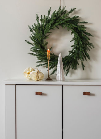 Christmas decor of the living room - a Christmas tree wreath on the wall, a brass candlestick with a burning candle, Christmas paper toys on a white chest of drawersの写真素材