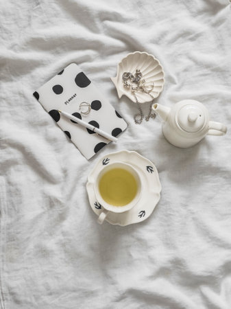 Cozy morning - a cup of green tea, a glider, women's jewelry on white sheets, top viewの写真素材