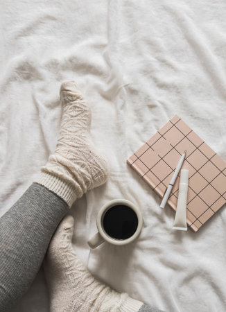 Women's legs in ribbed leggings, warm socks, coffee mug, diary on the bed, top viewの写真素材