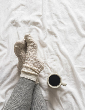Coffee break. Women's legs in leggings, warm socks and a mug of coffee on white sheets in bedの写真素材