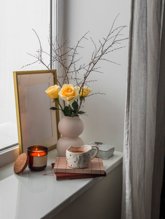 Cozy composition still life on the window - a bouquet of yellow roses, a cup of tea, books, a burning candle - a cozy house conceptの写真素材