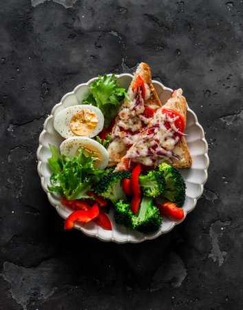 Delicious diet lunch, breakfast - boiled egg, fresh vegetables, broccoli, sweet pepper, lettuce and hot sandwiches with tomato and cheese on a dark background, top viewの写真素材