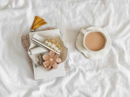 A cozy morning in bed - a cup of tea with milk, a bowl of cream, a hair clip, mascara, notebooks on white sheets in bedの写真素材