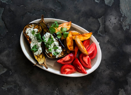 Grilled eggplant with tzadziki sauce, baked potatoes, fresh tomatoes on a dark background, top viewの写真素材