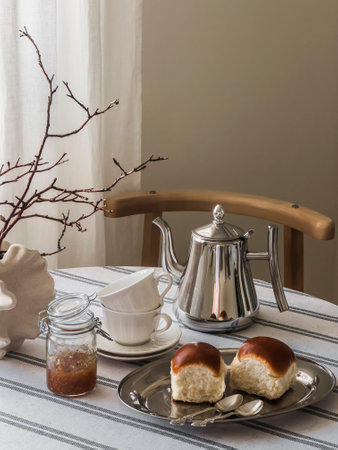 Homemade cozy tea party - brioche buns, fig jam, cups, teapot, vase on a round table with a tablecloth in the living roomの写真素材