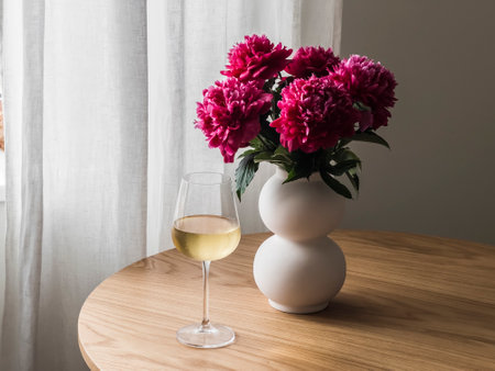 A glass of white wine, a bouquet of red peonies in a ceramic vase on a round wooden tableの写真素材