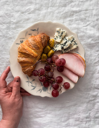 Delicious breakfast, snack - a woman's hand holds a plate with croissant, ham, grapes, blue cheese...