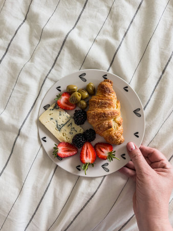 A woman's hand holds a plate with breakfast, snack - croissant, gorgonzola cheese, berriesの写真素材