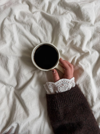 A hand in a brown knit sweater with lace detail holds a cup of black coffee against white bedding,...