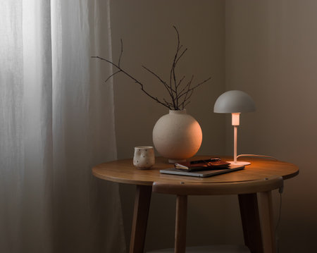 A cozy evening still life - a vase with branches, a lit table lamp, a laptop, a notebook, a glass of coffee on a round wooden table in the living roomの写真素材