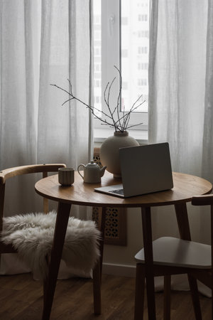 The cozy interior of the living room - a round wooden table with a ceramic vase, a laptop, a cup, and minimalist chairs by the windowの写真素材