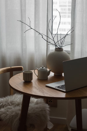 The cozy interior of the living room - a ceramic vase with branches, a laptop, a cup with coffee on a wooden table near the windowの写真素材