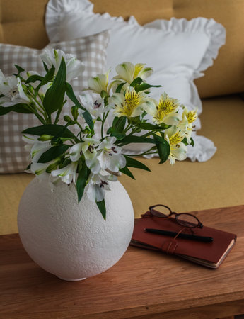 A bouquet of fresh flowers in a ceramic vase, a notebook on a wooden table near a sofa with decorative pillowsの写真素材