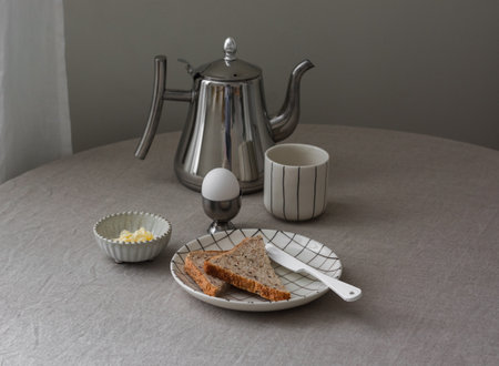 Breakfast served - boiled egg, whole grain bread, butter, coffee pot on the table with a gray tableclothの写真素材
