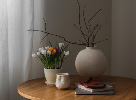 Cozy spring home still life - crocuses in a pot, a round ceramic vase, a laptop, a cup of coffee on a round wooden tableの写真素材