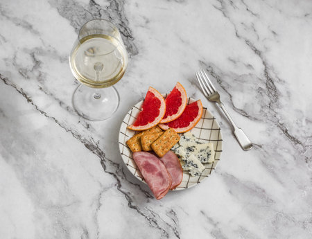 Aperitif on a marble background - glass of white wine, ham, blue cheese, cracker, grapefruit slicesの写真素材