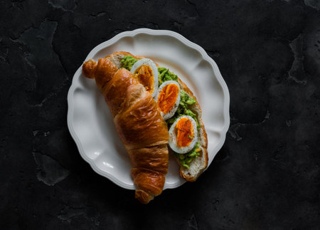 Croissant sandwich with avocado and boiled egg on a white ceramic plate on a dark background, top viewの写真素材