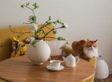 The cozy interior of the living room includes a round wooden table with a vase of flowers, a cup of tea, and a ginger cat. A cozy home conceptの写真素材