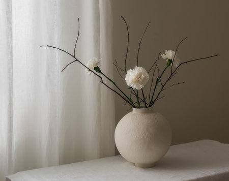 A composition of white carnations and dried branches in a ceramic round vase on a white tableの写真素材