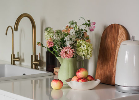 Cozy kitchen interior - a bouquet of summer flowers in a ceramic jug, a kettle, a bowl with apples, a cutting board on a stone table near the sinkの写真素材