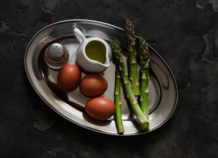Breakfast ingredients - fresh asparagus, eggs, olive oil, pepper on a metal plate, top viewの写真素材