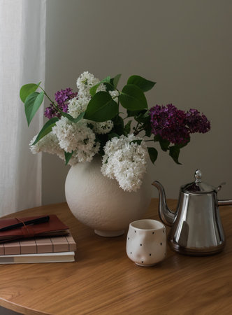 Cozy interior - a bouquet of lilacs, a coffee pot, a glass, books on a round wooden table in the living roomの写真素材