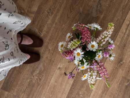 A bouquet of garden flowers in a vase and a woman's feet in suede shoes and a muslin dress on a wooden floorの写真素材