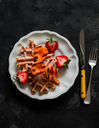 Traditional Belgian waffles with caramelized apricots and strawberries - a delicious dessert on a dark background, top viewの写真素材