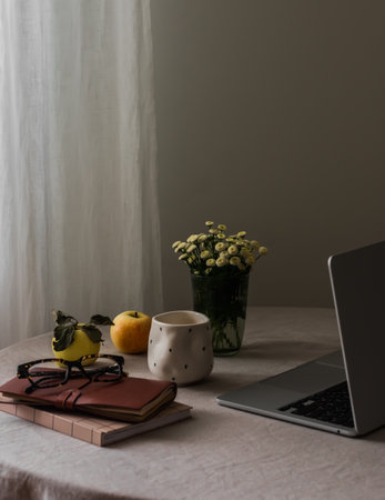 A cozy home interior - a laptop, books, a glass, apples, flowers on a round table with a gray linen...