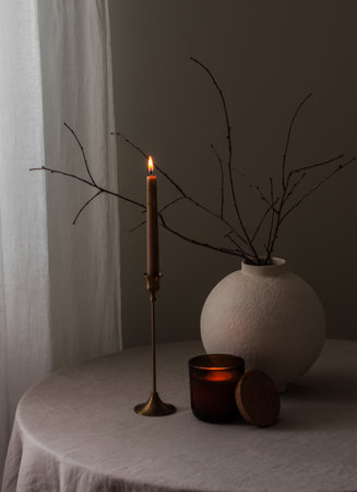 A cozy home still life - lighted candles, a ceramic vase with dry branches on a round table in the living roomの写真素材