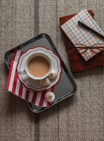 A cup of tea with milk, meringue on a metal tray, notebooks, glasses on the carpet, top viewの写真素材