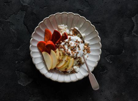 Delicious breakfast - cottage cheese with yogurt, apple, persimmon and granola on a dark background, top viewの写真素材
