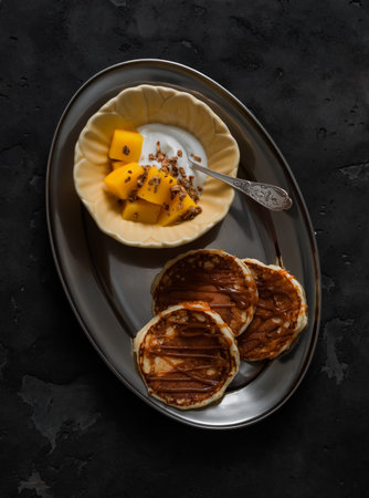 Delicious breakfast, brunch - pancakes with caramel sauce, yogurt with granola and mango on a dark...