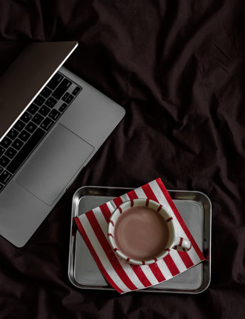 A cup of cocoa on a metal tray, a laptop on brown linens, top view. Lazy morning