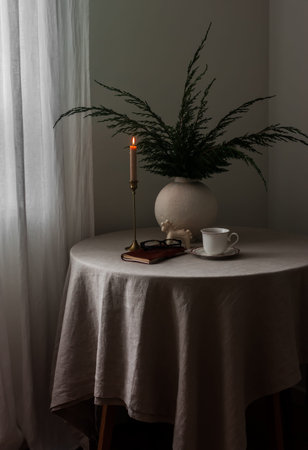 A round table with a gray tablecloth, a vase with juniper branches, a candle, and a cup of coffee in...