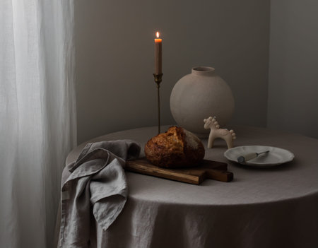 Sourdough bread, a lit candle, a vase, a plate on a round table with a gray tablecloth in the living...