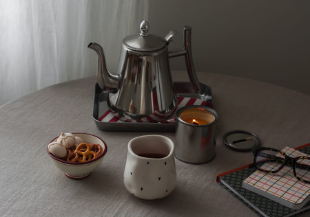 Metal teapot, cup, candle, biscuits, book on the table in the living roomの写真素材