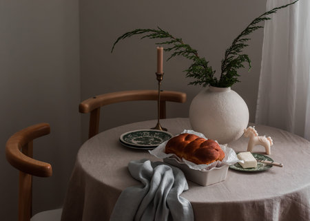 Cozy interior - freshly baked brioche, a vase with juniper branches, candles, dishes, butter on a...