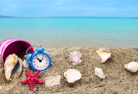 Shells,clock and bucket on sandy beach with sea in the backgroundの写真素材